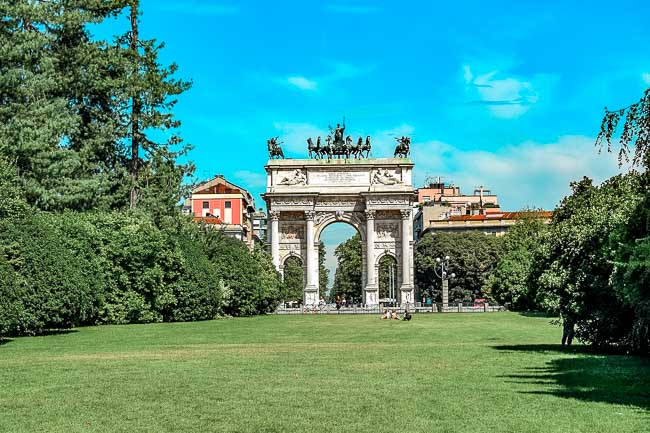 The famous archway with a statue of a chariot on top in Milan’s Parco Sempione with green grass in the foreground and a blue sky. Relaxing in the park is one of the best things to do in Milan.