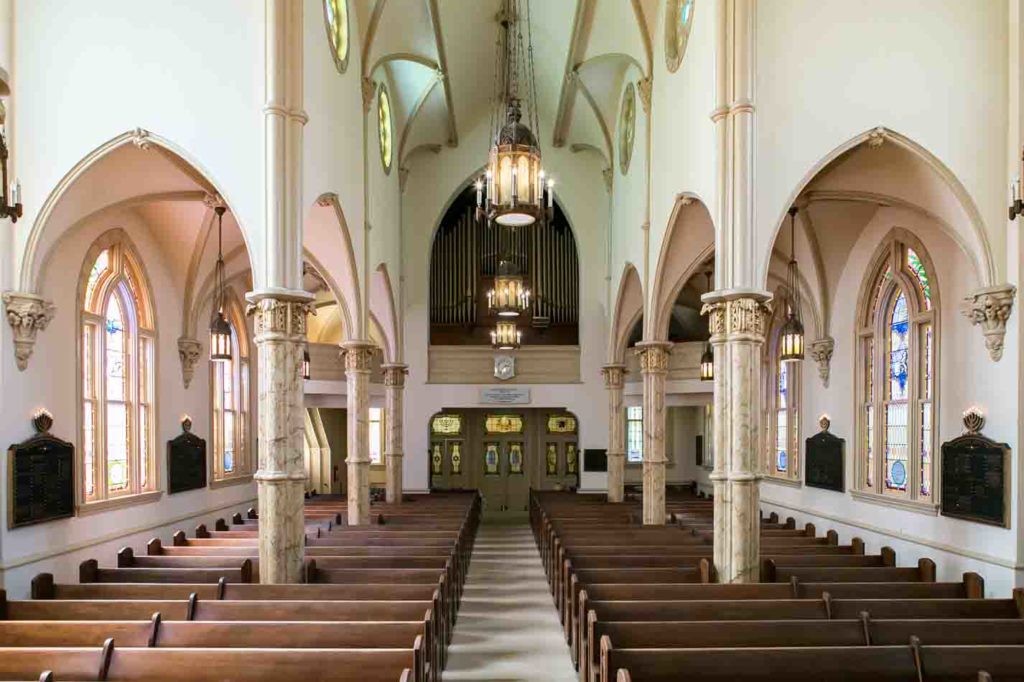 Interior of Congregation Mikveh Israel Savannah Ga, archways and columns fill the scene along with long rows of brown pews and stained glass windows along the sides. 