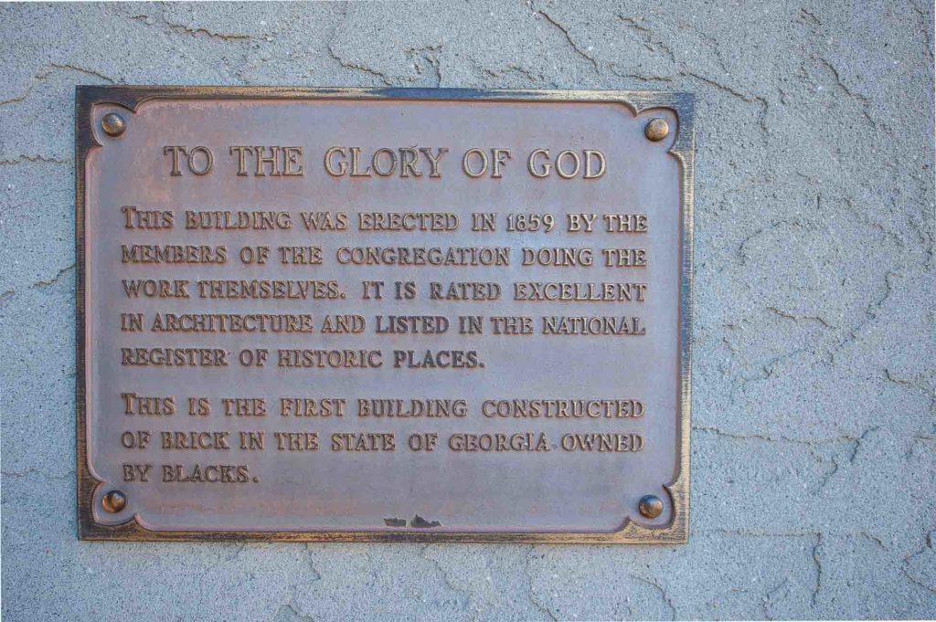 A bronze plaque mounted on a textured gray wall commemorates the First African Baptist Church of Savannah. The plaque reads: "To the Glory of God. This building was erected in 1859 by the members of the congregation doing the work themselves. It is rated excellent in architecture and listed in the National Register of Historic Places. This is the first building constructed of brick in the state of Georgia owned by Blacks." This site is an important part of Savannah historical sites. 
