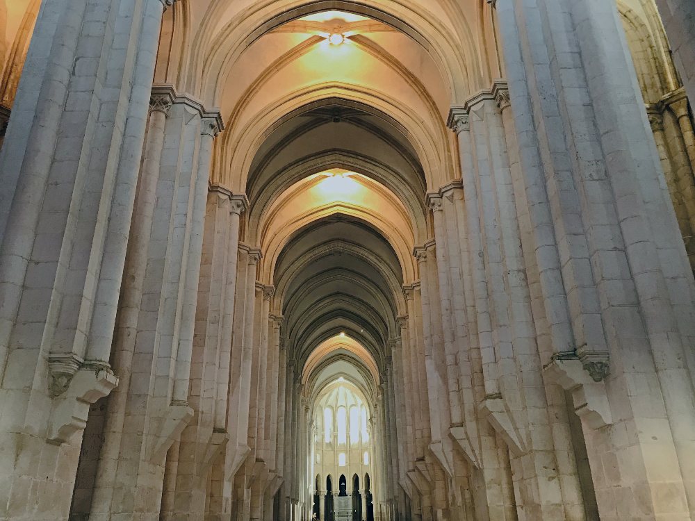 In the interior of the Cathedral at Alcobaça. Plain columns rise to a high arch and repeat through a long nave of a church towards the altar. 