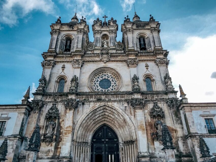 The facade of Alcobaça with three square towers and a large portal against a blue sky with clouds. The imposing building has one big rose window in the center. 