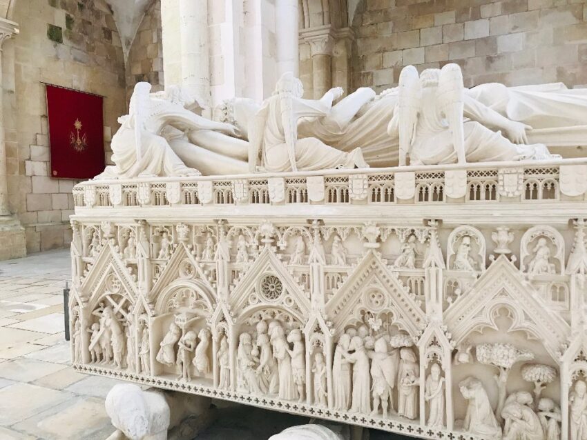 The tomb of Pedro and Ines at Alcobaça with a bright red flag in the corner. An intricate white marble tomb has the statues (laying down) of  Pedro and Ines, angels attending them, and very detailed scenes from their life carved into the side. 