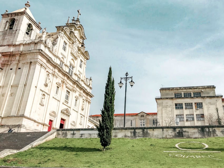Facade of the Sé Nova or New Cathedral in Coimbra Portugal all white with green grass in front with "Coimbra" mowed into the grass and a single thin green pine tree. 