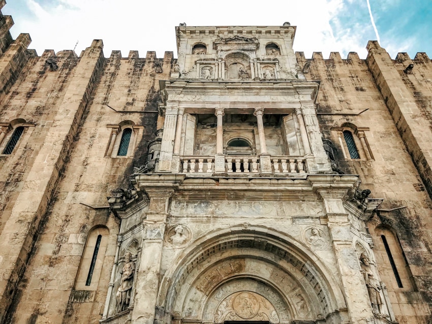 The Renaissance portal of the Old Cathedral of Coimbra. It is built so it juts out a bit with a side archway and then a square balcony with three pillars. The stone is a different color to the Cathedral surrounding it. 