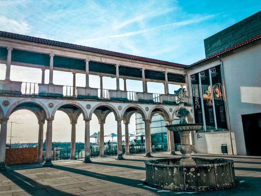 A double layer of see-through arches with a museum to the right. Through the arches you can see a balcony and a view over Coimbra rooftops. 