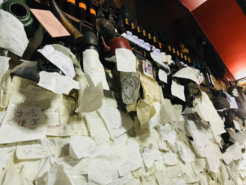 A wall with some kettled hung at the top and a red ceiling and hundreds of torn pieces of paper taped to the wall with notes and drawings completely covering the wall. 
