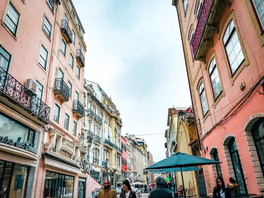 View from a street with a red buildling on the right and other buildings on the left, umbrellas and shops line the street.