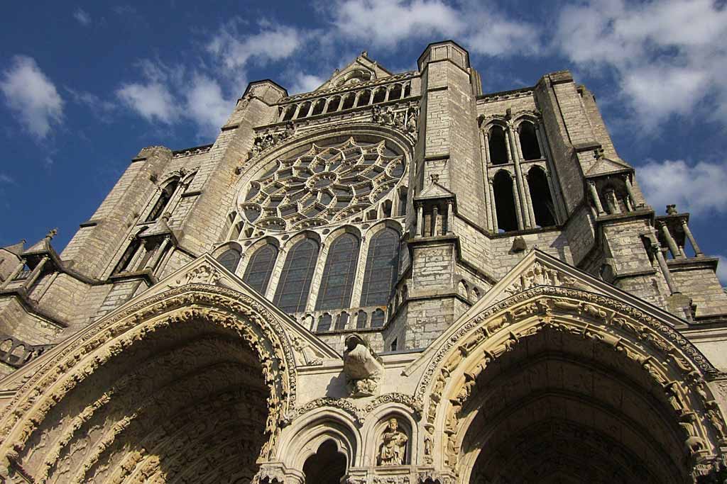 Facade of Chartres Cathedral with two vaulted arches and towers against a blue sky with clouds. 