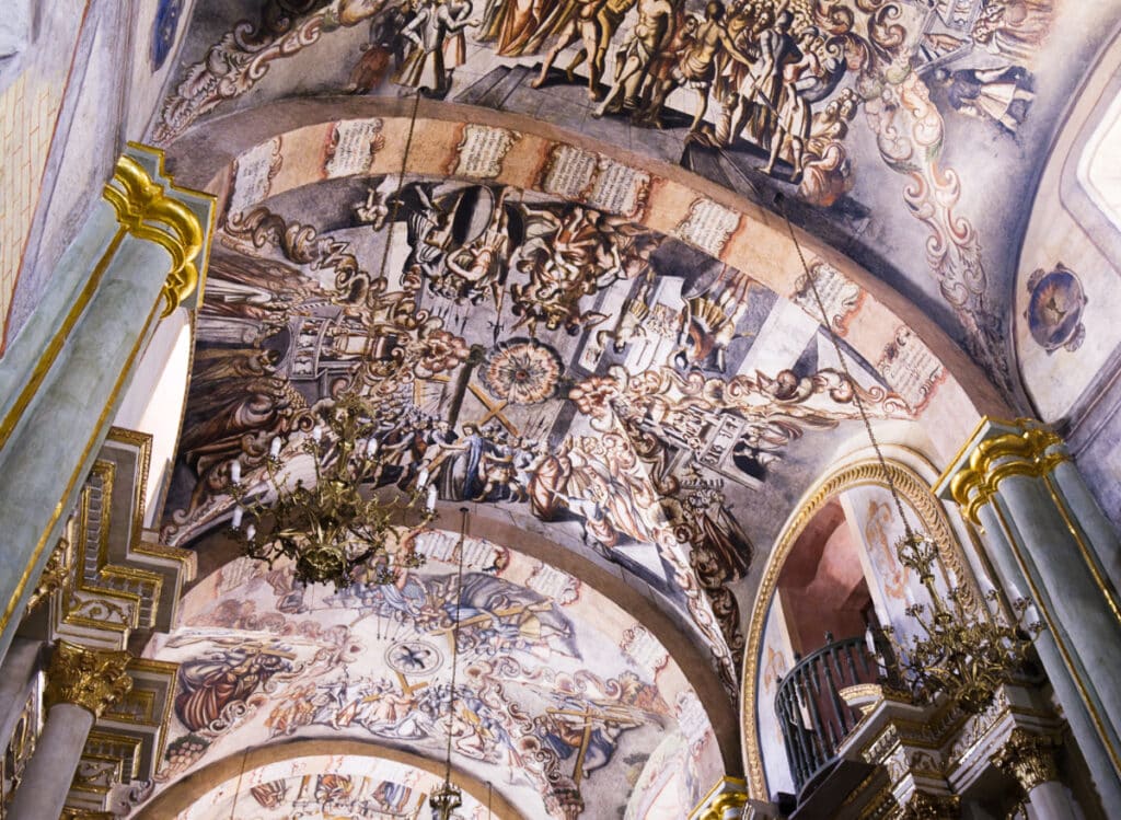 Grey columns with gilded gold caps lead to arched ceilings covered in a series of murals at the Sanctuary of Atotonilco, a pilgrimage site in Mexico. The vivid murals depict the life and death of Christ in mostly muted colors of red and ochre.