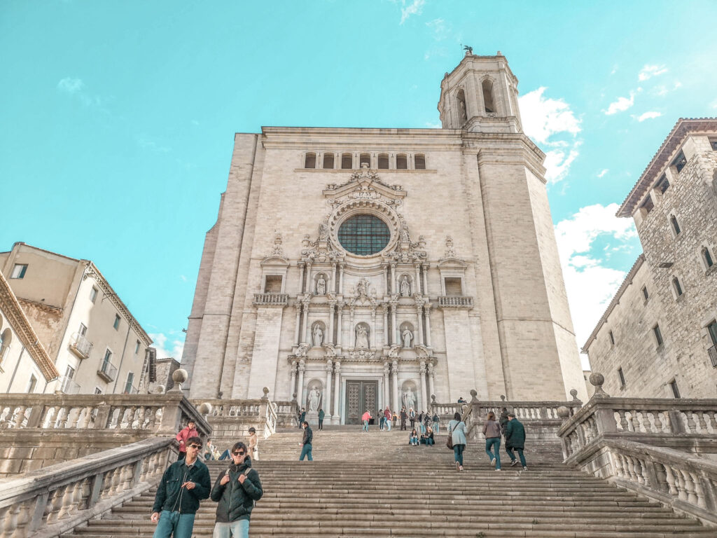 Looking up the steps to the Girona cathedral which has one bell tower and a big rose window over an arched portico with multiple niches. The sky is bright blue and there are building surrounding the cathedral.
