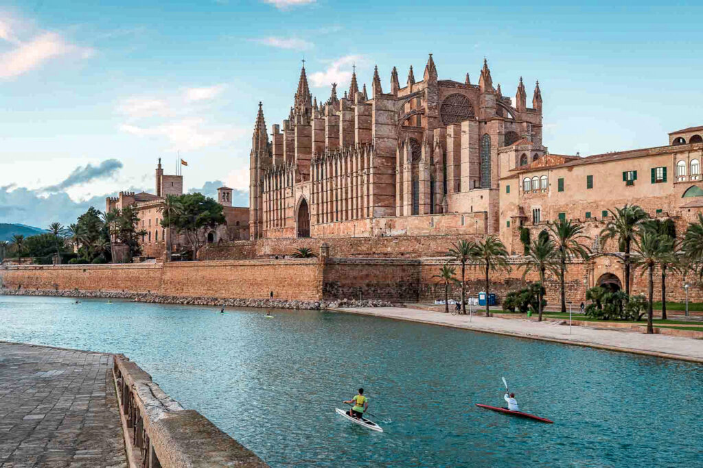 The medeival Palma Cathedral across a moat with kayakers against a blue sky. There are palm trees in the foreground the spires of the Palma Cathedral evoke a middle-eastern feel on the imposing edifice.