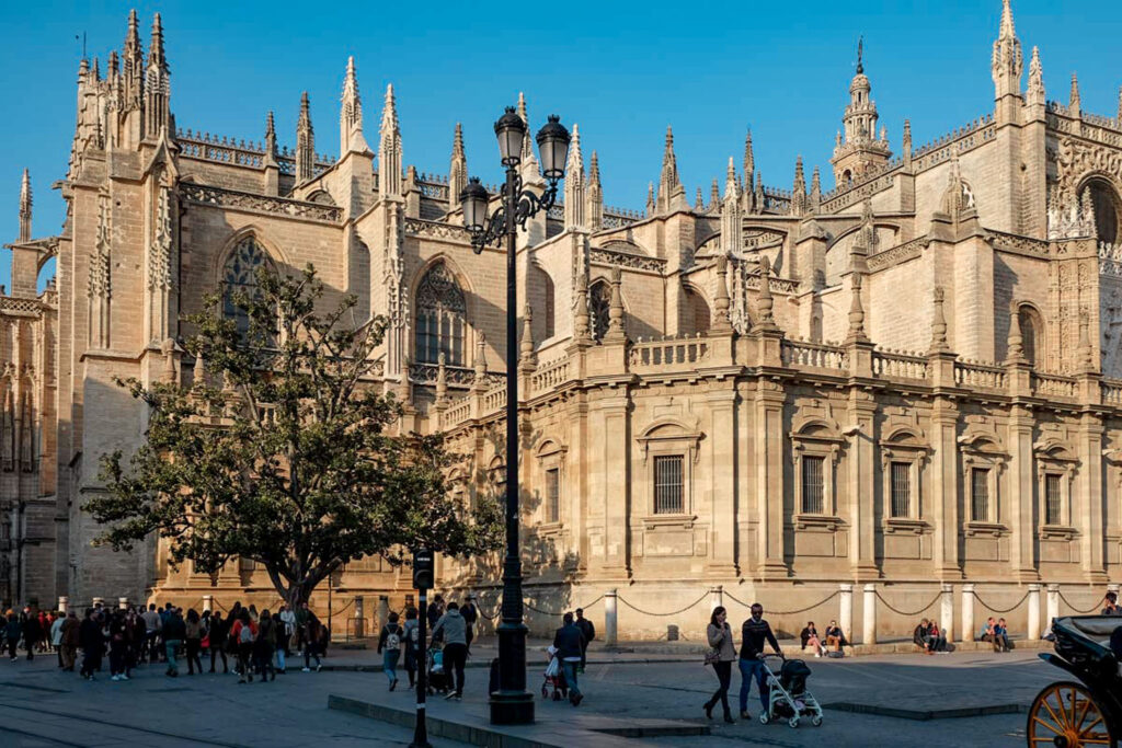 Seville Cathedral on a sunny day, one of the most famous cathedrals in Spain. A large Gothic cathedral in yellow stone with lots of ornate spires, arched windows and a tall bell tower. The cathedral is on a square with people walking below.
