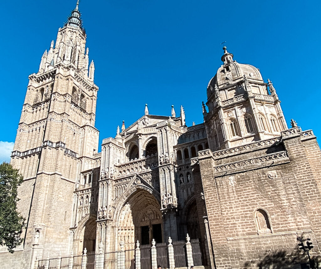 A striking image showcasing the ornate gothic architecture of Toledo Cathedral in Spain, featuring its towering spire and intricate facades against a bright blue sky.