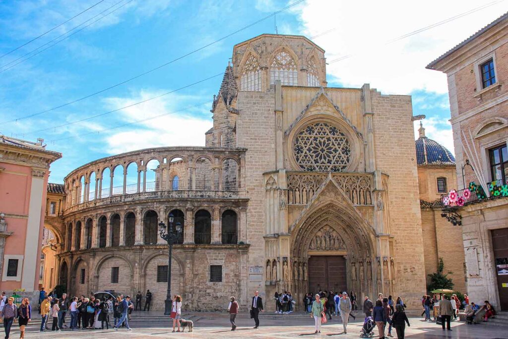 Valencia Cathedral - A towering Gothic and Romanesque cathedral with intricate architecture in shades of beige and brown. The sun casts a warm glow on the facade, highlighting the ornate details. A crowd gathers in the plaza below.