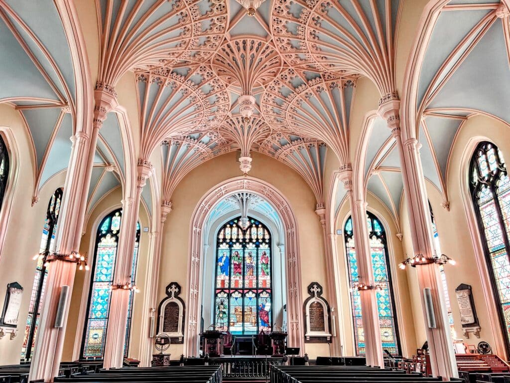 The interior of the Unitarian church in Charleston, SC, showcases a striking Gothic revival design. The high, arching ceilings are supported by slender columns, all painted in a light peach hue. The intricate tracery and fan vaulting above are delicate and ornate. Stained glass windows cast colorful light into the space, illuminating the wooden pews and the central aisle leading to a modest altar. 