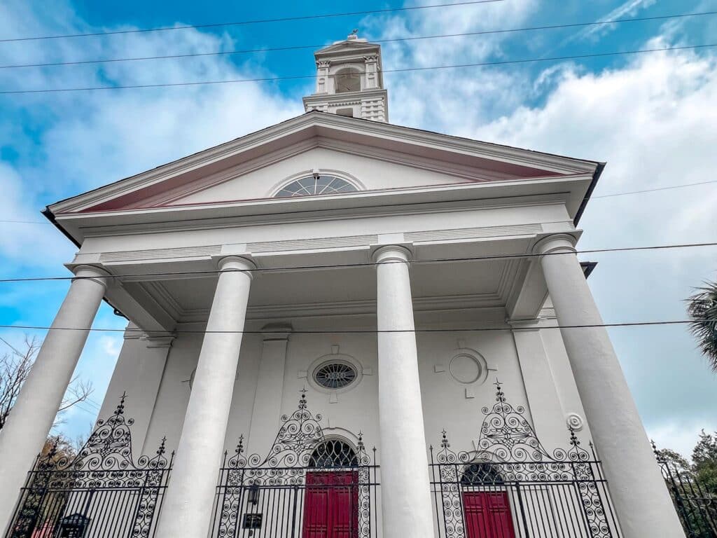 This image depicts the front of a neoclassical church, the Cathedral of St. Luke and St. Paul in Charleston with a pristine white facade and towering columns. The architecture includes a prominent pediment supported by large, round columns, under which sits a vibrant red door. Circular and oval windows add decorative elements to the structure. The foreground features an elaborate wrought-iron gate. Above, the sky is partly cloudy