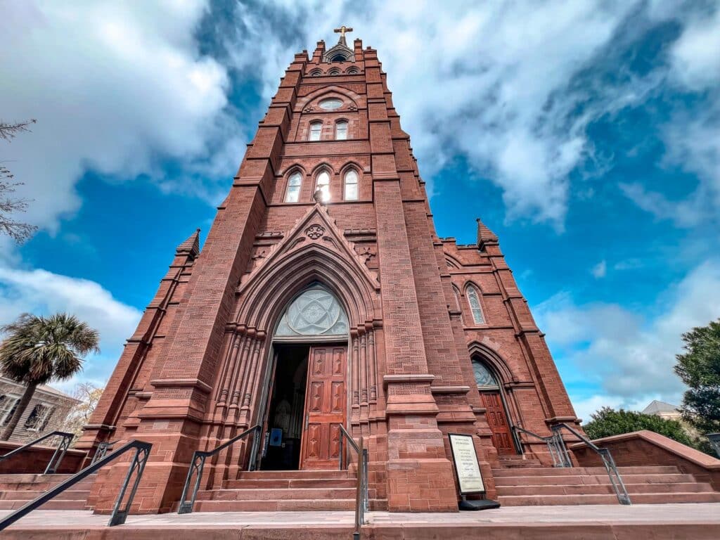 The front entrance of the Cathedral of St. John the Baptist in Charleston, south Carolina in red brick against a blue sky with three large pointed doors. This is one of the most famous churches in Charleston, SC. 