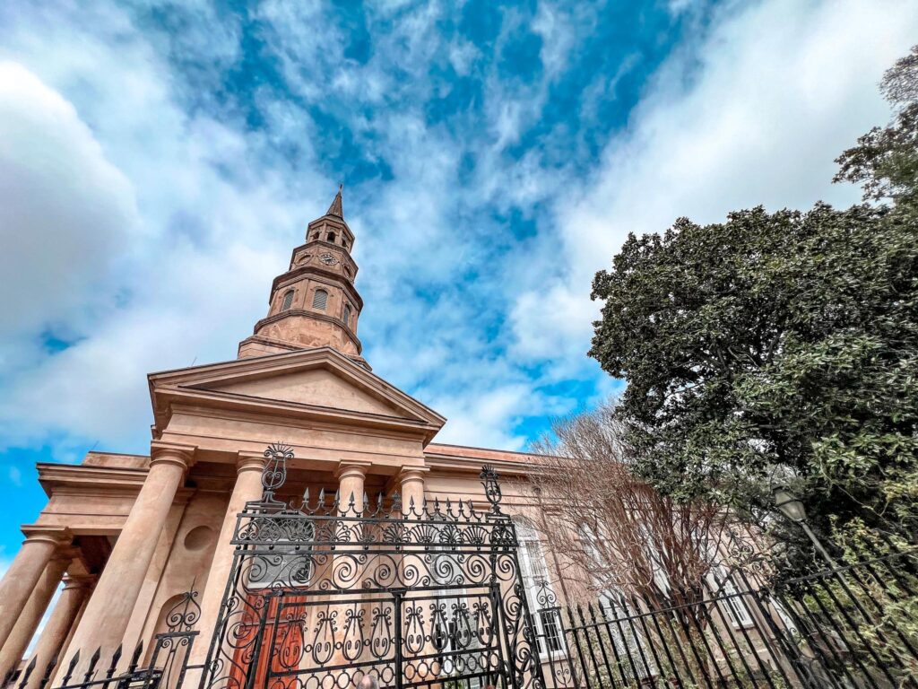 The photograph shows the Cathedral of St. Luke and St. Paul in Charleston, one of the most stately churches in Charleston with a grand beige facade and a towering steeple piercing a sky filled with wispy clouds. The church's architecture includes classical columns and a pediment that echoes traditional design. A decorative wrought-iron gate encloses the foreground. Green foliage from mature trees peek from the sides.