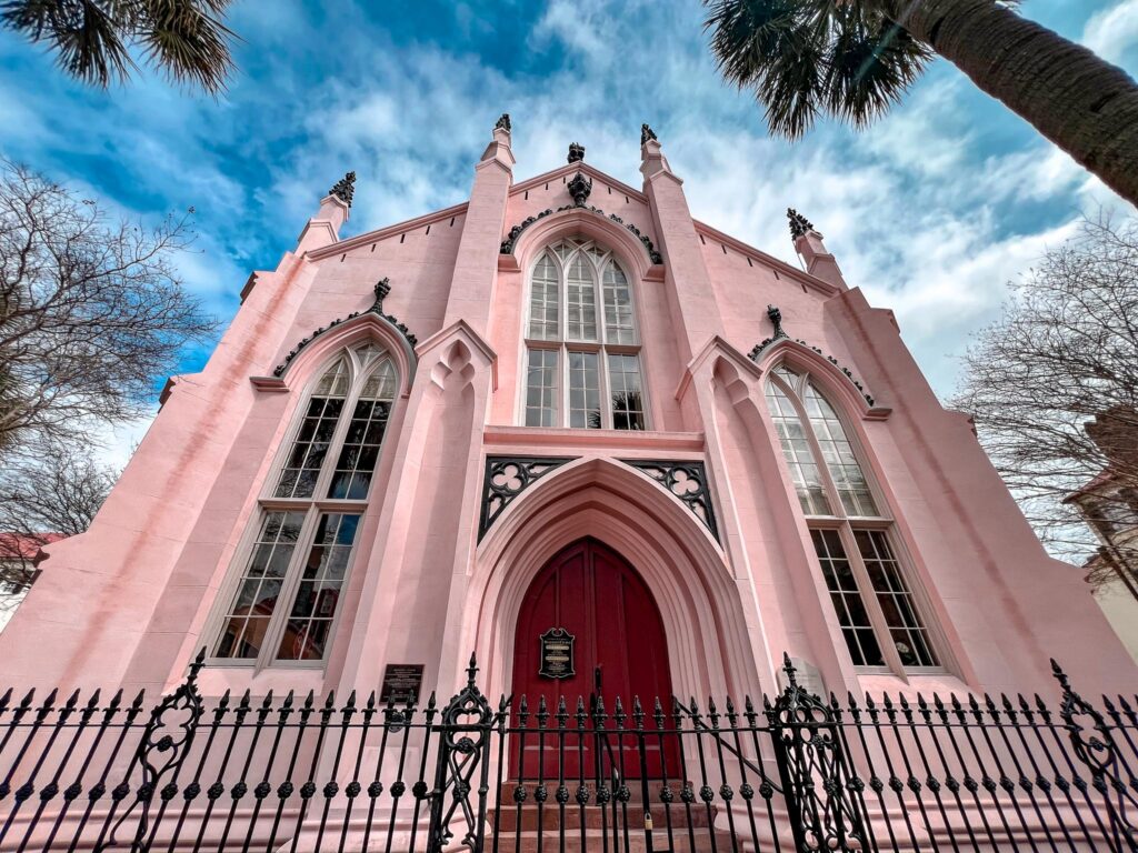 A view of the French Hugenot Church in Charleston, a pink Gothic revival church in Charleston, featuring a series of pointed arches and intricate window tracery. The church's facade is accentuated by little sharp spires and decorative finials atop each pinnacle. The main entrance has a bold red door set within a deeply recessed arch. A black wrought-iron fence surrounds the property. Above, the sky is clear with a few wispy clouds.