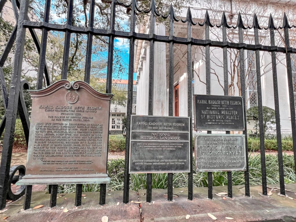 The image features several historical plaques mounted on an ornate black wrought-iron fence. The plaques provide information about Kahal Kadosh Beth Elohim, identifying it as both a National Historic Landmark and a site on the National Register of Historic Places. The text commemorates the establishment of the synagogue in Charleston in 1749, highlighting its significance in the history of American Judaism. Visible in the background is the white facade of the synagogue, partially obscured by the fence and framed by mature trees and clear skies.