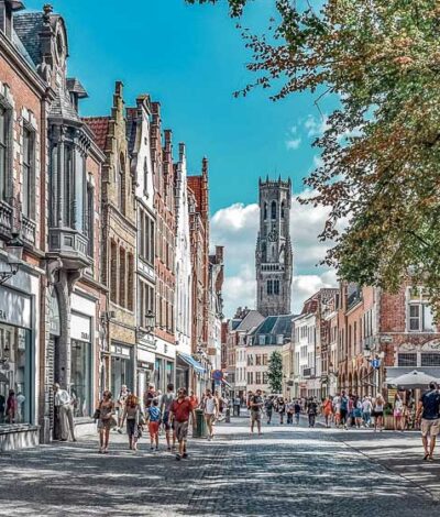 A street scene in Bruges, featuring historic buildings with ornate facades lining a cobblestone street. Tourists stroll beneath trees, with the iconic Bruges Belfry tower visible in the distance against a clear blue sky. The image captures the essence of a bustling summer day in this medieval Belgian city that is worth spending one day in Bruges.