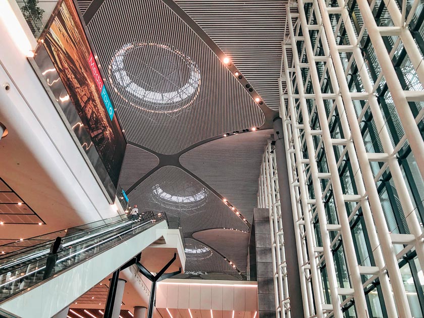 The bustling IST Istanbul Airport has beautiful architecture. The phoeo shows an escalator to the left, and geometric and dome patterns on the ceiling and a large decorative glass wall to the right.