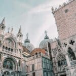Architectural details of St. Mark's Basilica in Venice. Ornate Gothic and Byzantine facades with arches, domes, and spires. Intricate stone carvings and sculptures adorn the building. Pink-tinted marble walls contrast with white stone elements. Two large domes visible in the background. Cloudy sky above.