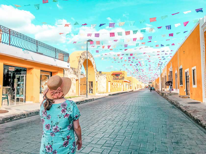 Cate with a floral shirt and hat standing in a street in Mexico with yellow houses and flags flying across them.