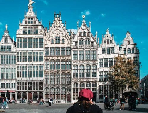 A person is taking a picture of four guild buildings in Antwerp Belgium with a blue sky. The buildings are highly intricate and come to a point and the one on the left is tallest. Enjoying architecture is one of the best things to do in Antwerp, Belgium