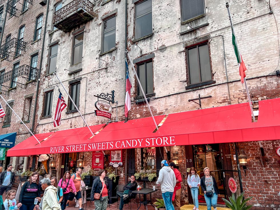 A view of River Street Sweets Candy Store in Savannah with a bright red awning and flags coming out of it. There are lots of people milling about on busy River Street, one of the best historical places to visit in Savannah and best places to eat in Savannah. 