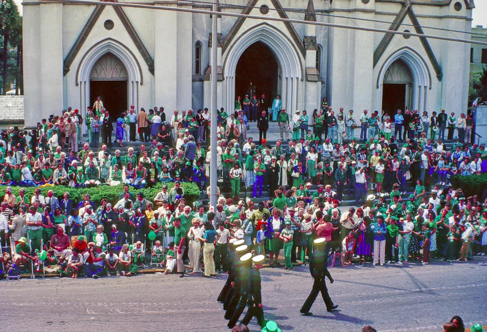 A parade marches by in front of the Savannah Cathedral. Hundreds of people are crowded in front to watch all wearing green. The photo has a grainy historic look and was likely taken in the 80s. 