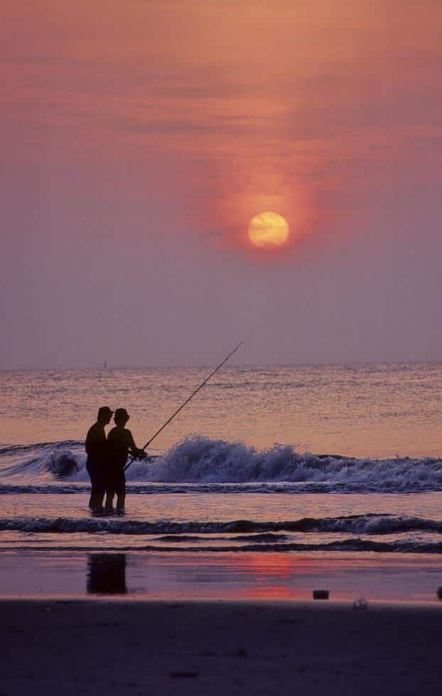 Through a misty sunset, two people are fishing on the beach on Tybee Island. One holds a very long fishing pole. A wave crashes near them. The whole photo is bathed in misty, red light. 